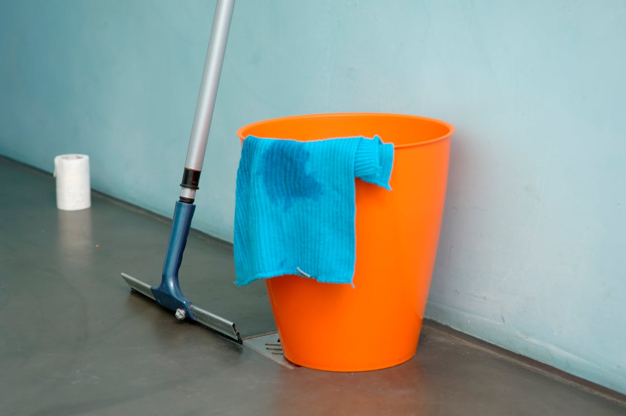services-03 Close-up of cleaning supplies including orange bucket, mop, and blue rag indoors.